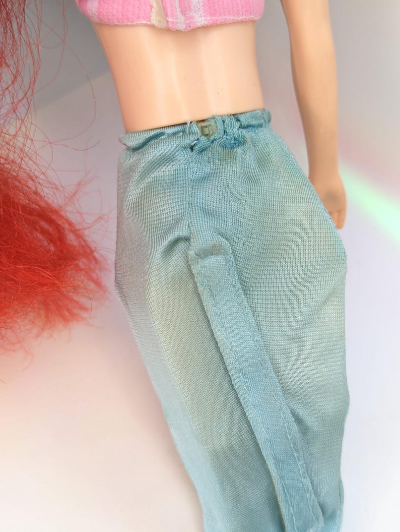 Op de afbeelding: Een pop met een roze en wit gestreept topje en een lichtblauwe broek. De pop heeft rood haar en een lichte huidskleur. De broek heeft een kleine knoopsluiting in de taille en een verticale naad.