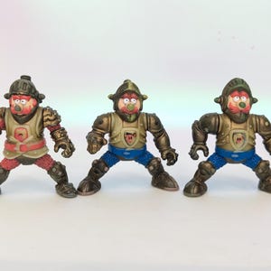May include: Three toy figures of cartoon knights in armor. The figures have red faces, helmets, and armor in shades of brown, gold, and red. They are posed with their legs spread and arms out.