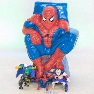May include: A large blue and red plastic Spider-Man storage container shaped like the character sitting down, accompanied by four small action figures including a green and purple villain, a black creature, a small Spider-Man, and a white skeleton figure.