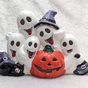 May include: A ceramic Halloween decoration featuring a group of smiling white ghosts, a carved orange pumpkin, and a black cat wearing a witch hat. The ghosts have black eyes and red mouths. The pumpkin has a green stem.