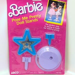 May include: A Barbie Pose Me Pretty Doll Stands package. The package is pink with a blue star-shaped stand and a purple circular stand. The text on the package reads "Barbie" and "Pose Me Pretty Doll Stands."