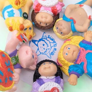 May include: A collection of vintage Cabbage Patch Kids dolls. The dolls have various hair colors and outfits, including a doll holding a heart-shaped sign that reads "I Love You". A blue and white drawing of a child is visible.