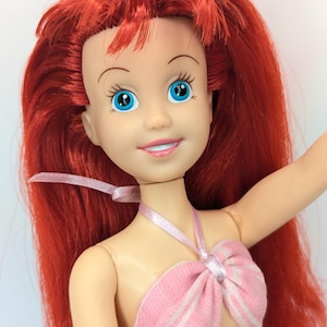 Op de afbeelding: Pop met rood haar en blauwe ogen, gekleed in een roze en wit gestreepte top. De pop heeft een lachende uitdrukking en een roze lint om haar nek. Het haar van de pop is lang en vloeiend.
