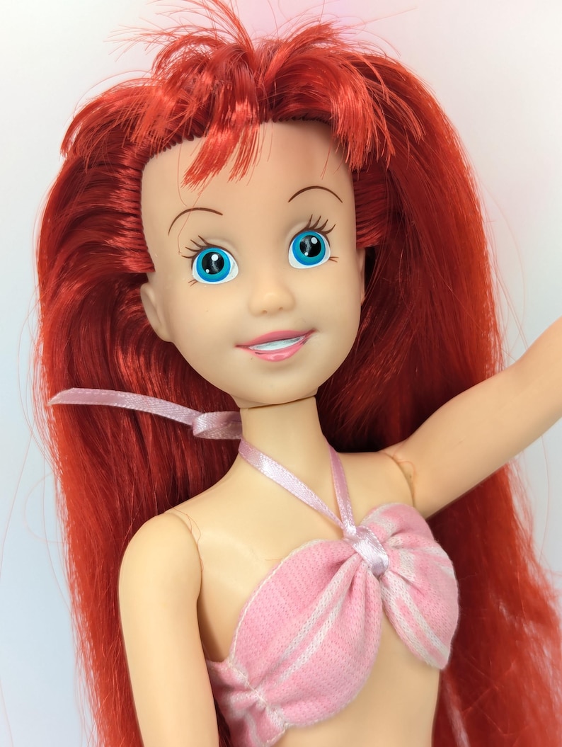 Op de afbeelding: Pop met rood haar en blauwe ogen, gekleed in een roze en wit gestreepte top. De pop heeft een lachende uitdrukking en een roze lint om haar nek. Het haar van de pop is lang en vloeiend.