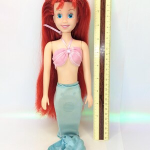 Op de afbeelding: Een pop van een zeemeermin met lang rood haar, blauwe ogen en een roze top. De pop heeft een lichtblauwe zeemeerminstaart en staat naast een liniaal die tot 30 cm meet.