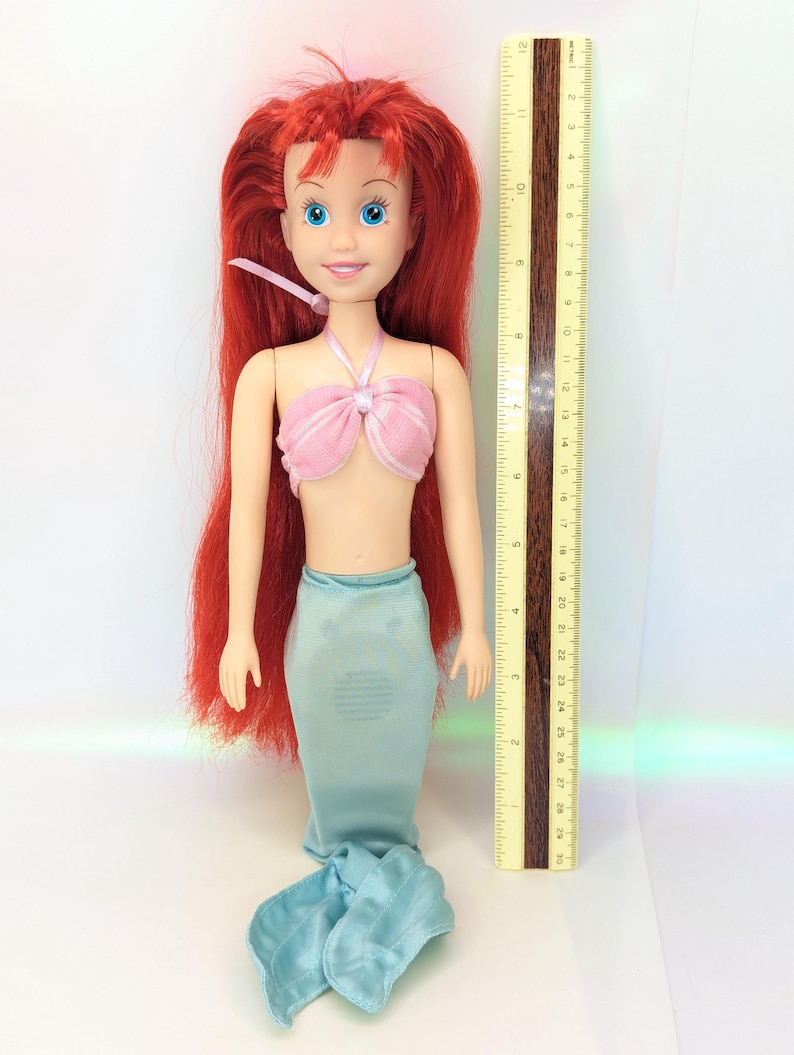 Op de afbeelding: Een pop van een zeemeermin met lang rood haar, blauwe ogen en een roze top. De pop heeft een lichtblauwe zeemeerminstaart en staat naast een liniaal die tot 30 cm meet.