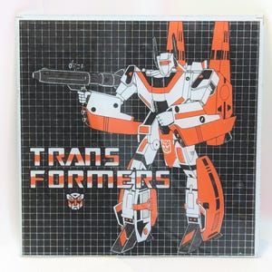 May include: A vintage Transformers graphic featuring a robot character in white and orange, holding a weapon. The image is set against a black grid background, with the word "TRANSFORMERS" prominently displayed.