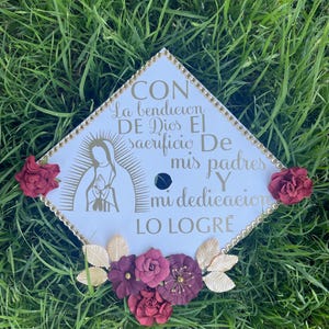 Op de afbeelding: Een witte afstudeerhoed versierd met gouden glitter en een gouden afbeelding van de Maagd Maria. De hoed heeft ook een bordeauxrode bloemversiering en de tekst "CON la bendicion DE Dios El sacrificio De mis padres Y mi dedicacion LO LOGRE".