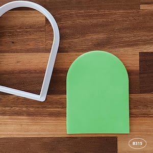 May include: A green fondant cutout in the shape of an arch, next to a white plastic cookie cutter in the same shape. The background is a wooden surface.
