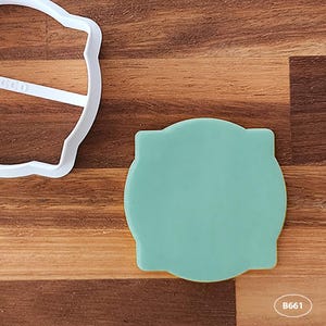 May include: A light green plastic cookie cutter shaped like a circle with rounded edges. The cookie cutter is on a wooden surface.
