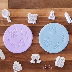 May include: A set of 10 white plastic cookie cutters in the shape of birthday party decorations. The cutters are arranged on a wooden surface with two purple and blue silicone mats. The mats have the same shapes as the cutters, but in a different arrangement. The text "B712" is visible on the wooden surface.