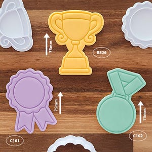 May include: A selection of colourful plastic cookie cutters in various shapes, including a yellow trophy, a purple ribbon, and a green medal. Each cutter is approximately 7.5 cm in height. The cutters are arranged on a wooden surface.