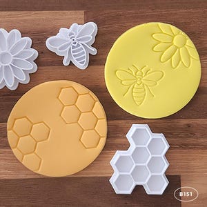 May include: A set of four white plastic cookie cutters in the shape of a bee, a daisy, a honeycomb, and a honeycomb pattern. The cutters are on a wooden surface with two yellow and orange fondant circles that have been embossed with the bee, daisy, and honeycomb patterns.