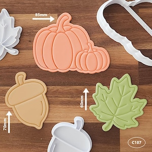 May include: A collection of autumn-themed cookie cutters and stamps including a peach-coloured double pumpkin shape measuring 85mm, a tan acorn shape measuring 70mm, and a light green leaf shape measuring 60mm, alongside white plastic outlines.