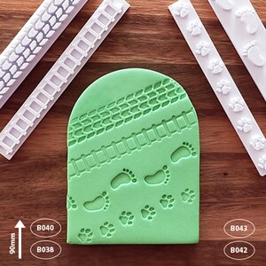 May include: Four white silicone cake decorating tools with different designs. The tools are on a wooden surface. The designs include tyre tracks, a fence, footprints, and paw prints. The tools are labelled B040, B038, B043, and B042.