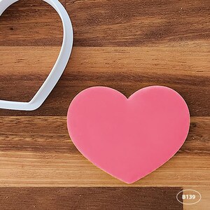 May include: A pink heart-shaped cookie cutter on a wooden surface. The cutter is made of white plastic and has a smooth finish.