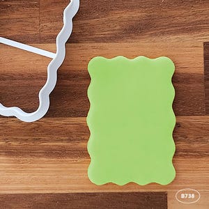 May include: A green, wavy-edged cookie cutter in the shape of a rectangle. The cutter is on a wooden surface.