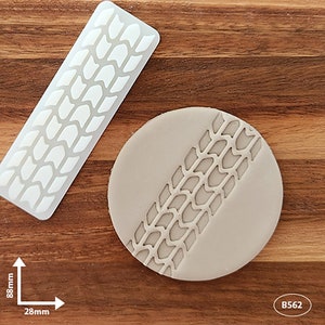 May include: A white silicone mould with a tyre tread pattern. The mould is 8.8 inches long and 2.8 inches wide. The mould is shown with a sample of the design imprinted in grey fondant.