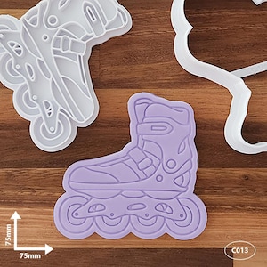 May include: Roller skate cookie cutters in white and lavender. The lavender cutter is 7.5cm x 7.5cm. The image shows the outline of a roller skate, with details of the wheels and boot. The white cutter is a roller skate outline as well. The image is on a wooden surface.