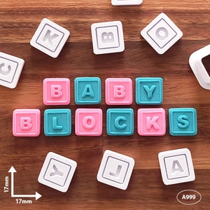 May include: A set of 10 white plastic alphabet blocks spelling out "BABY BLOCKS". The blocks are 1.7 cm square and feature raised letters. The blocks are arranged in a line on a wooden surface.