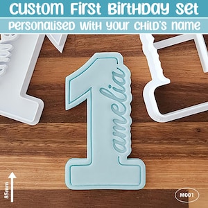 May include: A light blue number one cookie cutter with the name "amelia" in script. The text "CUSTOM First BIRTHDAY set" and "personalised with your child's name" is at the top. The cutter is 8.5cm in height.