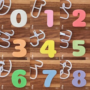 May include: A set of ten cookie cutters shaped like the numbers 0 through 9. The cutters are made of white plastic and are arranged in a grid pattern on a wooden surface. The numbers are in different colours, including pink, orange, yellow, green, and purple.