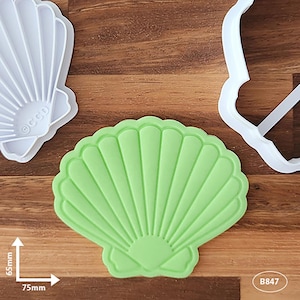 May include: Green plastic seashell cookie cutter with a detailed design. The cutter is 7cm wide and 6.5cm tall.  The cutter is shown next to a white plastic seashell cookie cutter.