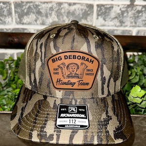 May include: A camouflage trucker hat with a brown leather patch that says "Big Deborah Hunting Team" with a cartoon image of a child holding a hunting bow and arrow. The patch also says "Hunt Hard" and "Snack Harder". The hat is adjustable and has a Richardson logo.