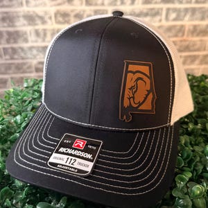 Könnte beinhalten: Schwarz-weiße Trucker-Cap mit einem Lederaufnäher mit dem Bundesstaat Alabama und einem Elefanten-Design. Die Cap hat einen gebogenen Schirm mit weißen Nähten und eine Mesh-Rückseite. Das Etikett der Marke Richardson ist sichtbar.