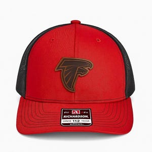 Può includere: Cappellino da camionista rosso e nero con una toppa in pelle marrone con un logo di uccello stilizzato. Il cappellino ha una visiera curva e un retro in rete. L'etichetta Richardson 112 è visibile.
