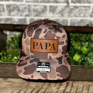 May include: A brown and tan camouflage trucker hat with a brown leather patch that says "PAPA" on the front. The hat is adjustable and has a black mesh back.