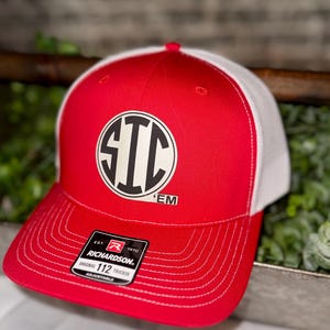 May include: Red and white trucker hat with a circular logo featuring the letters "STC" in black. The hat has a curved brim and a mesh back. The tag reads "RICHARDSON ORIGINAL 112 TRUCKER ADJUSTABLE".