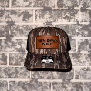 Könnte beinhalten: Trucker-Cap mit Camouflage-Muster und braunem Lederaufnäher. Der Aufnäher trägt den weißen Schriftzug "THEM DAWGS IS HELL". Die Kappe hat einen schwarzen Mesh-Rücken und einen gebogenen Schirm. Das Etikett der Marke Richardson ist sichtbar.
