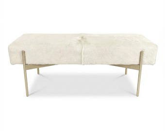 May include: A rectangular ottoman with a cream-colored cowhide top and gold-colored metal legs. The legs are angled and provide support to the upholstered top. The ottoman is a neutral color and would complement many interior design styles.