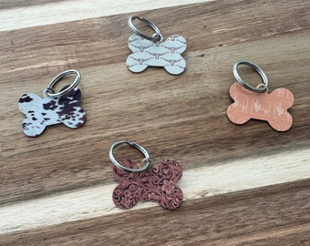 May include: Four dog bone-shaped keychains with different patterns. The keychains are made of metal and have a silver finish. The patterns include a cowhide print, a floral design, a cactus print, and a southwestern design.