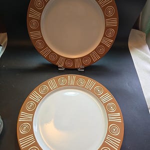 May include: Two ceramic plates with a brown rim featuring a repeating pattern of white circles and lines. The center of each plate is white. The plates are displayed on a dark surface.