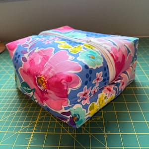 May include: A square, zippered pouch with a floral pattern. The fabric features large pink and smaller blue, yellow, and teal flowers on a blue background. The zipper is white, and the pouch is sitting on a green cutting mat.