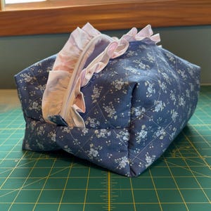 May include: A quilted, navy blue floral print bag with a light pink ruffled trim. The bag has a zipper closure and a diamond pattern. The bag is sitting on a green cutting mat.