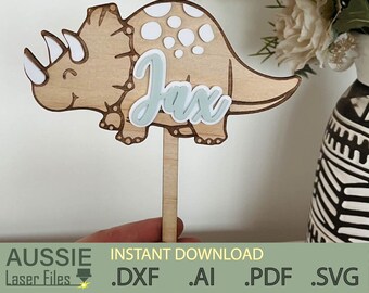 May include: A wooden triceratops dinosaur cake topper with the name "Jax" in light blue script. The dinosaur is light brown with white accents and a wooden stick for insertion. The bottom of the image has the text "AUSSIE Laser Files" and file type extensions.
