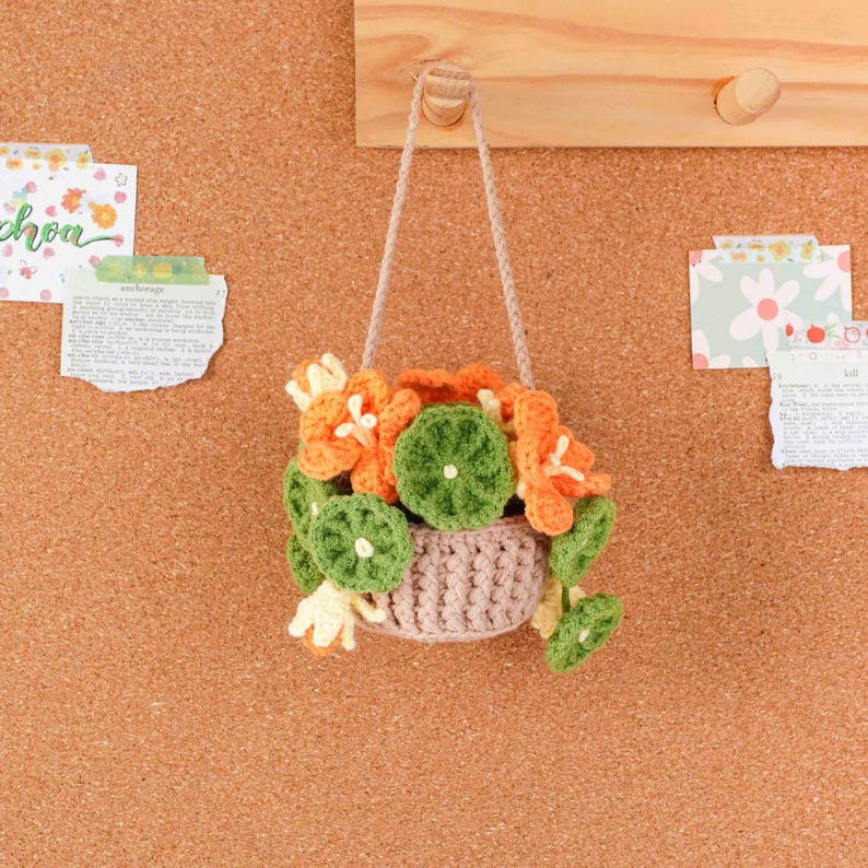 May include: A handmade crochet hanging basket filled with orange and green flowers. The basket is a light brown color and is suspended by a beige cord. The flowers are a mix of orange, green, and cream colors. The basket is hanging on a wooden board.