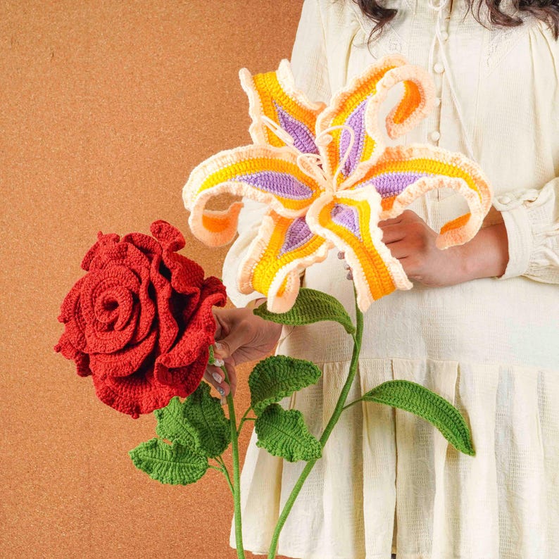 Op de afbeelding: Gehaakte bloemen: een levendige rode roos en een grote lelie met gele, oranje en paarse bloemblaadjes. De lelie heeft groene bladeren en een stengel. De roos is dieprood. Handgemaakte bloemen.