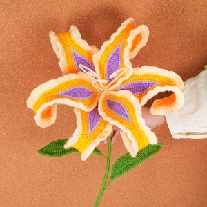 Op de afbeelding: Een grote, handgemaakte gehaakte bloem in tinten geel, oranje en paars. De bloem heeft gedetailleerde bloemblaadjes en groene bladeren, met een lange groene stengel. De bloem wordt door een persoon vastgehouden.