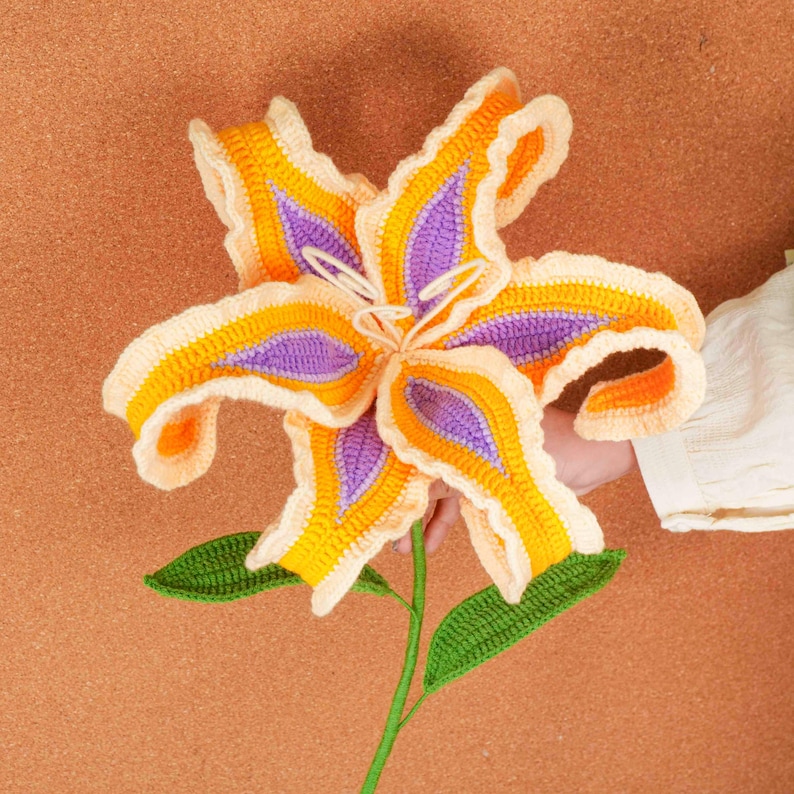 Op de afbeelding: Een grote, handgemaakte gehaakte bloem in tinten geel, oranje en paars. De bloem heeft gedetailleerde bloemblaadjes en groene bladeren, met een lange groene stengel. De bloem wordt door een persoon vastgehouden.