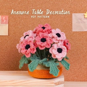 Könnte beinhalten: Eine gehäkelte Anemonen-Tischdekoration in einem kleinen orangefarbenen Topf. Die Blumen sind rosa mit schwarzen Zentren und grünen Blättern. Der Text "Anemone Table Decoration PDF Pattern" steht oben.