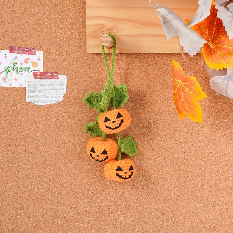 May include: A handmade Halloween decoration featuring three crocheted orange pumpkins with black jack-o'-lantern faces. The pumpkins are connected by green crocheted leaves and a green hanging string. The decoration is displayed against a corkboard background.