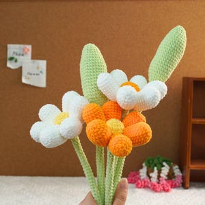 May include: A bouquet of crocheted flowers with white, orange, and yellow petals. The stems are green and the flowers are arranged in a hand.
