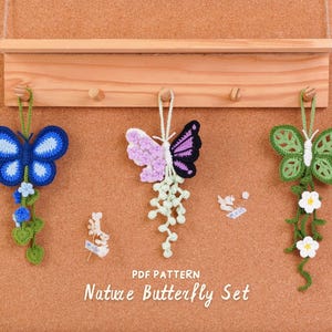 May include: Three crocheted butterfly ornaments hanging from a wooden rack. The left is blue with green vines, the center is purple and white with a dangling vine, and the right is green with white flowers. Text reads: PDF PATTERN Nature Butterfly Set.