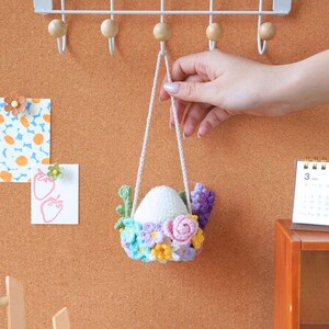 May include: A white crocheted egg with a floral wreath of pink, purple, blue, and yellow flowers hanging from a white string.
