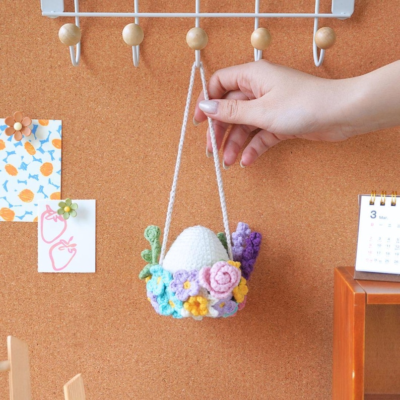 May include: A white crocheted egg with a floral wreath of pink, purple, blue, and yellow flowers hanging from a white string.