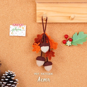 May include: A handmade crochet acorn ornament with brown, orange, and cream colors, hanging from a wooden peg. The ornament is accompanied by a decorative leaf and berries. The text "PDF PATTERN Acorn" is visible.